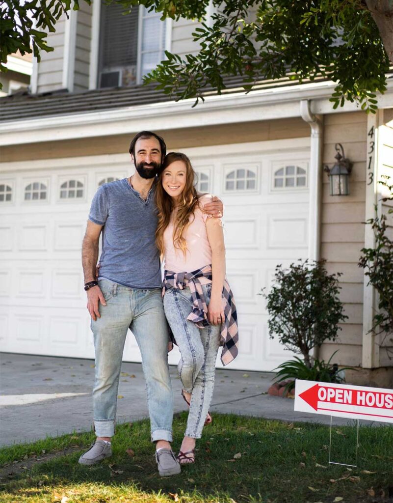A joyful couple standing in front of their new home with an open house sign, symbolizing the new beginnings and joy of homeownership.
