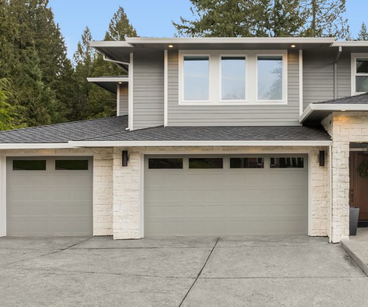 A modern home exterior featuring dual garages with white doors, stone detailing, and a sleek design against a backdrop of trees.